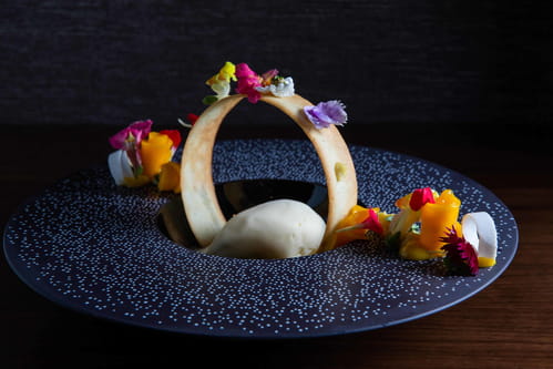 Halo high end dessert with vanilla tuile, coconut ice cream, passion fruit caramel, mango curd, and mango decorated with edible flowers on black disc speckled with sugar