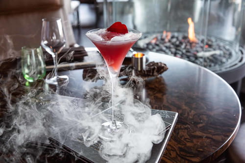 Craft cocktail with dry ice and fireplace in background at Bellevue steakhouse